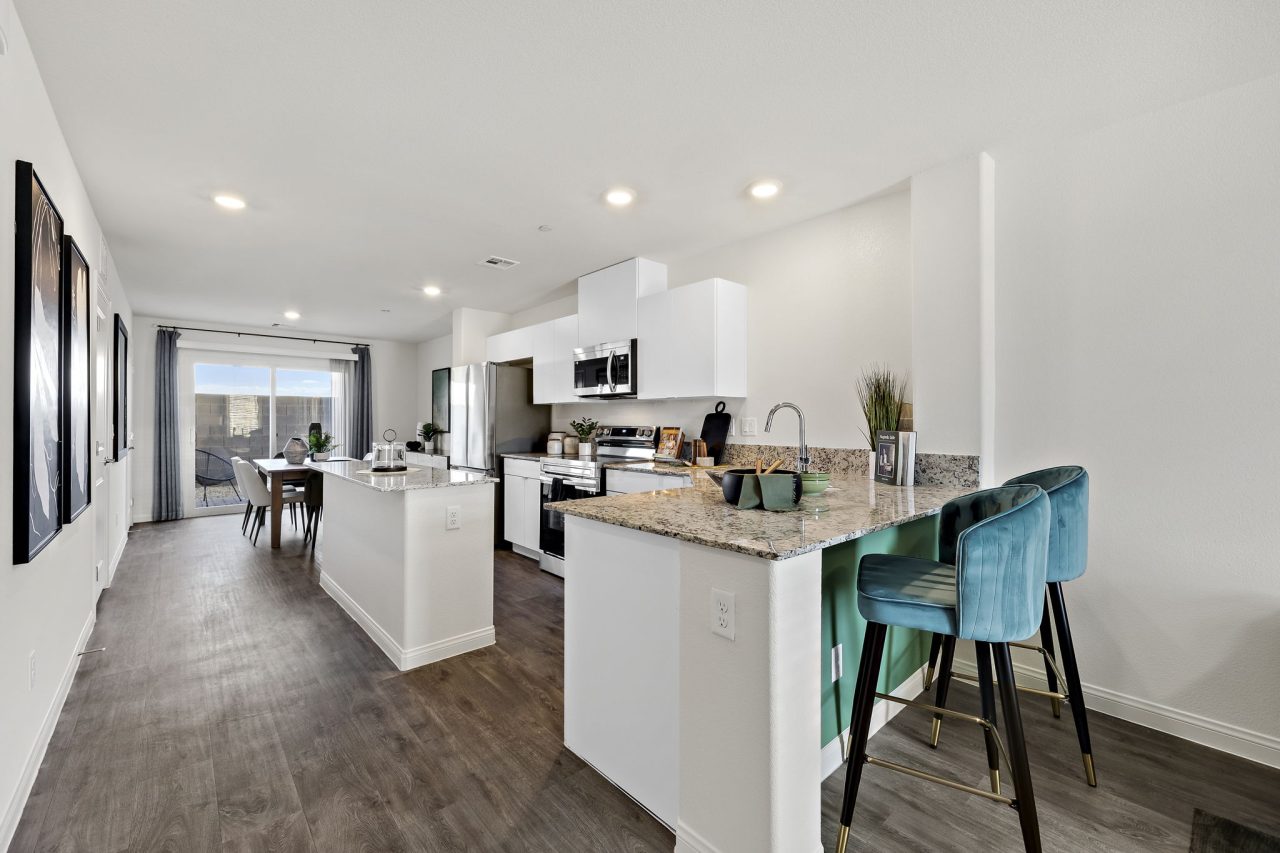 Modern open-concept kitchen featuring white shaker-style cabinets, granite countertops, a peninsula with upgraded vinyl flooring and teal velvet barstools in an Achieve model home.