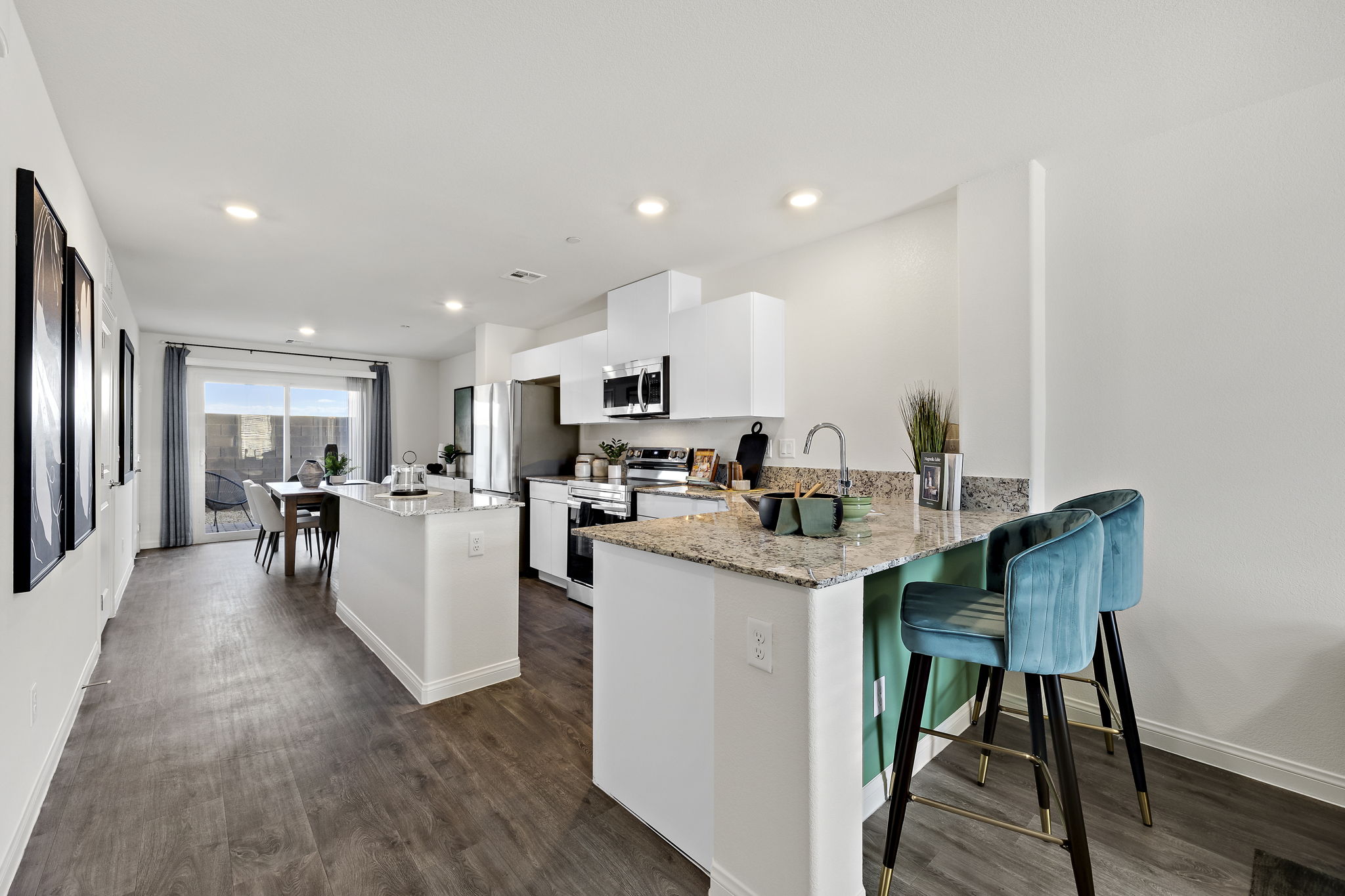Modern open-concept kitchen featuring white shaker-style cabinets, granite countertops, a peninsula with upgraded vinyl flooring and teal velvet barstools in an Achieve model home.