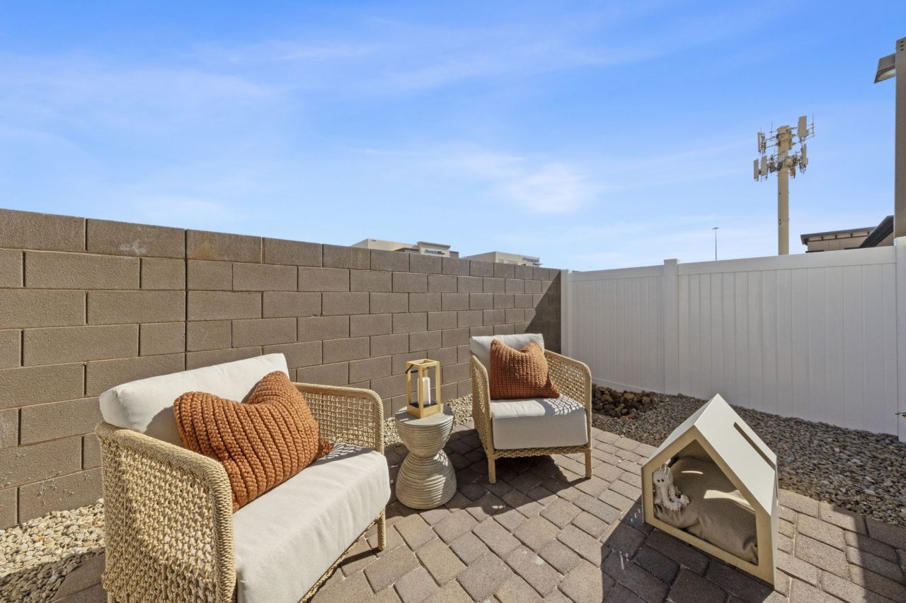 Private enclosed backyard patio featuring brick pavers, modern woven chairs with orange pillows, and a small dog house at a Solaris home.