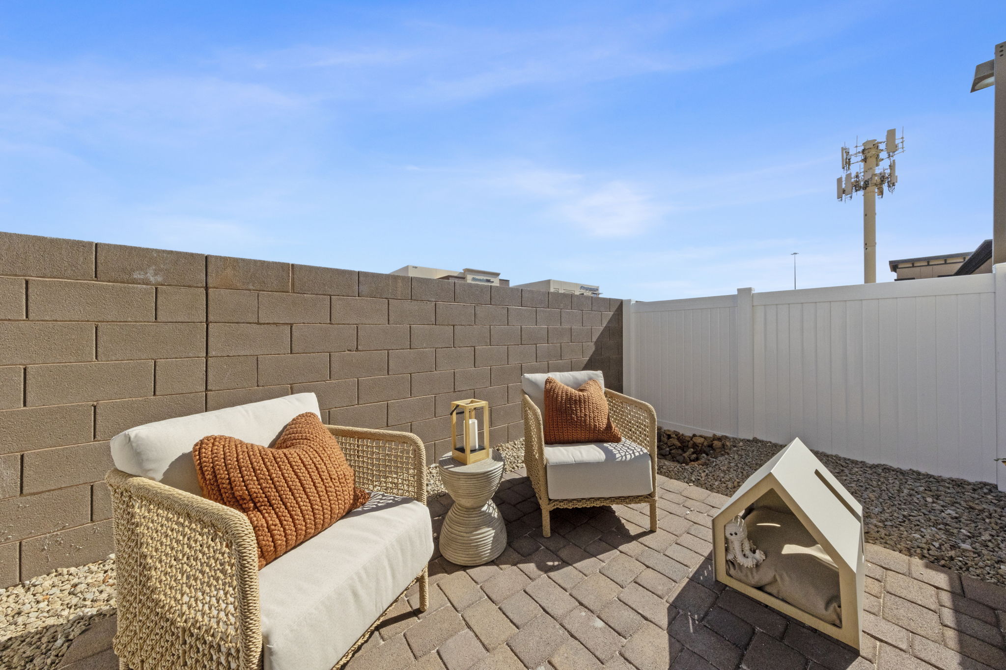 Private enclosed backyard patio featuring brick pavers, modern woven chairs with orange pillows, and a small dog house at a Solaris home.