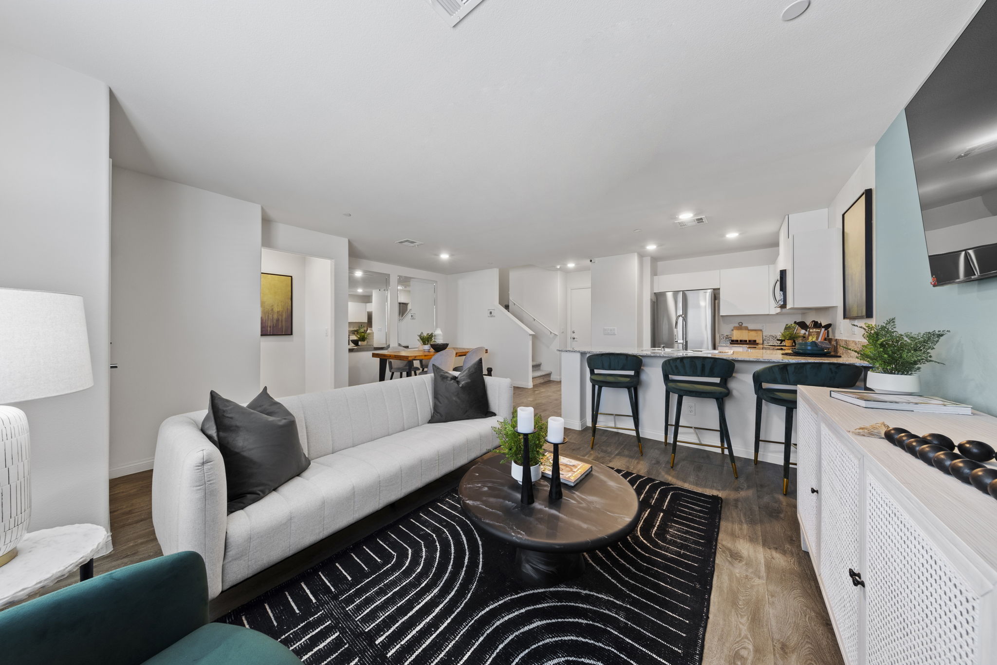Expansive open-concept living and kitchen area featuring upgraded vinyl flooring, a white modern sofa, and a kitchen island with teal barstools in a Prosperity model home.