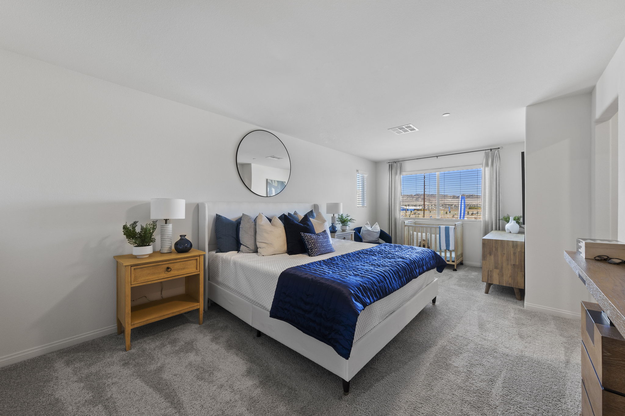 Spacious primary bedroom featuring a white upholstered bed with blue bedding, a large round mirror, and a crib near the window in a Prosperity model home.