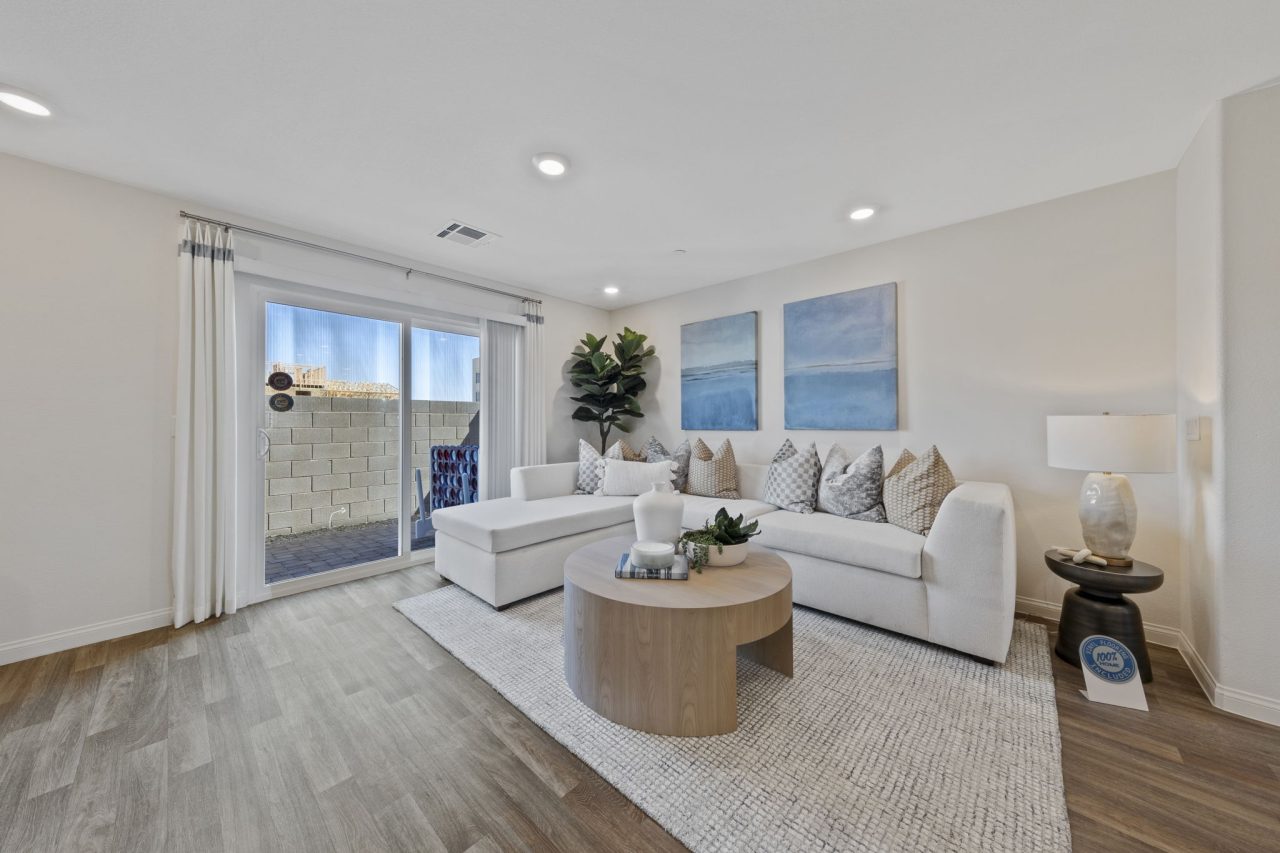 Bright living room featuring upgraded vinyl flooring, a white sectional sofa, a round wood coffee table, and blue abstract art in a Solaris model home.