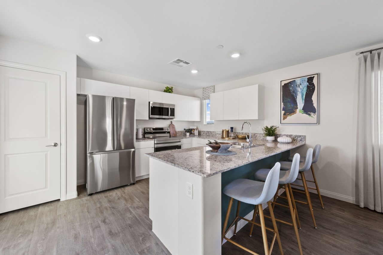 Modern open-concept kitchen featuring white cabinetry, granite countertops, upgraded vinyl flooring, and a peninsula with blue barstools in a Success model home.