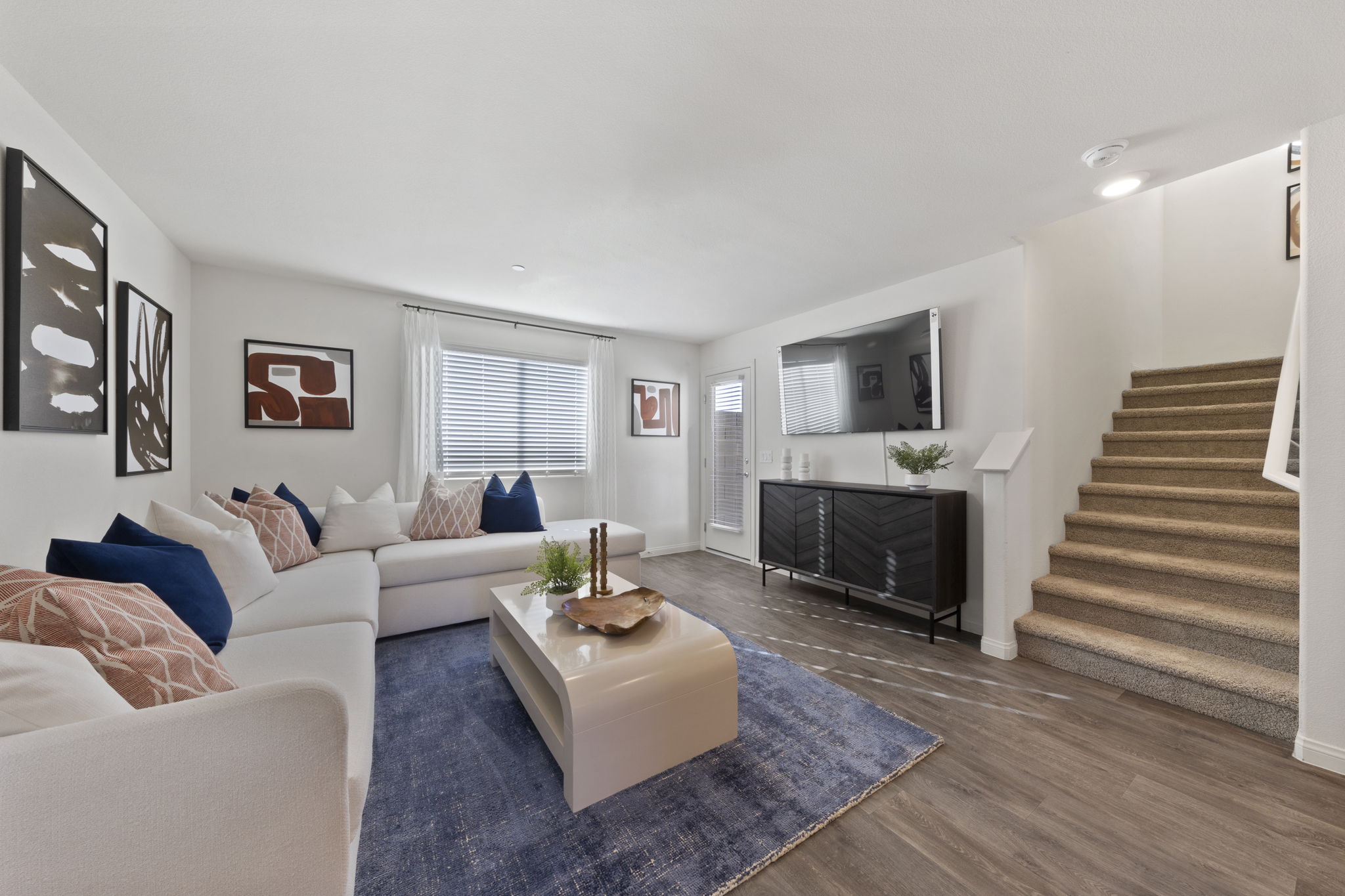 Bright living room featuring upgraded vinyl flooring, a large white sectional sofa, and carpeted stairs leading to the second floor in a Success model home.