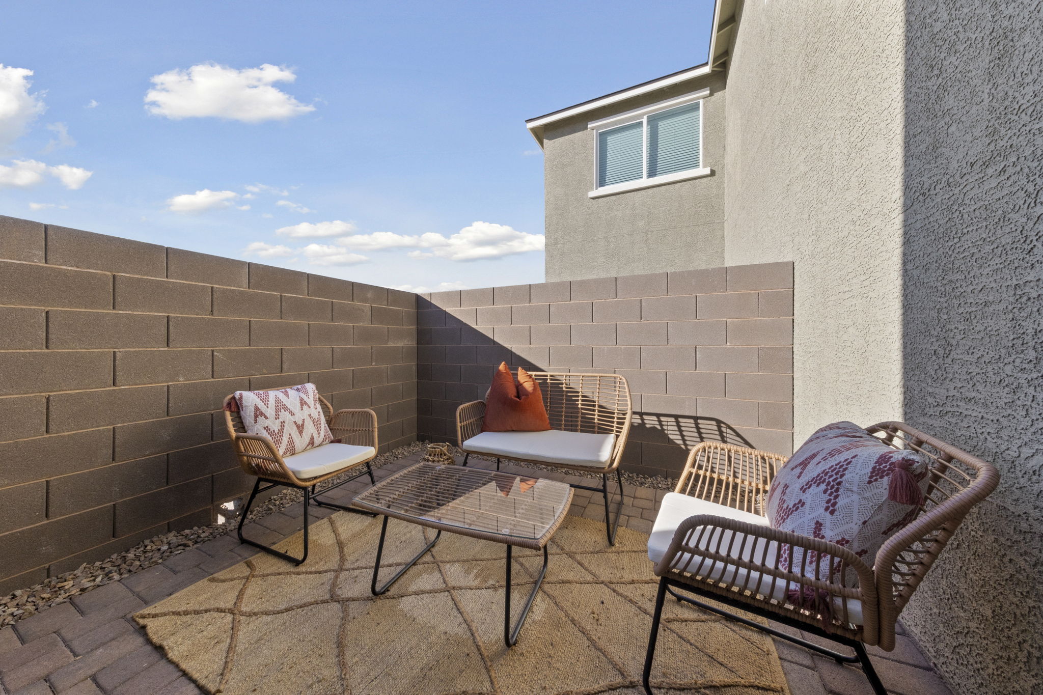 Cozy private patio area featuring brick pavers, a tan rug, and woven outdoor furniture with orange and patterned pillows at a Touchstone Living home.