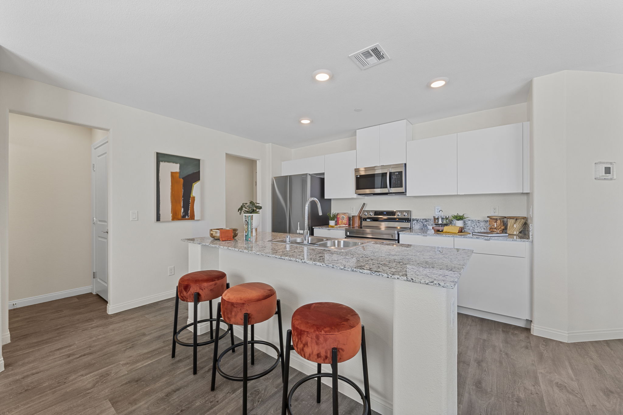Open-concept kitchen with white cabinetry, stainless steel Samsung® appliances, and a granite island with three orange barstools.