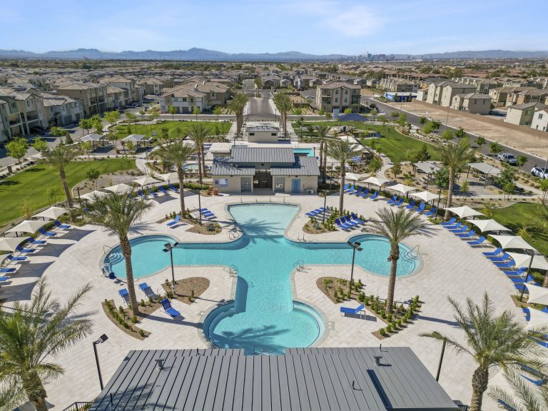 Aerial view looking down at the cross-shaped central swimming pool, clubhouses, and green parks at the Watercolor planned community, with the Las Vegas skyline in the distance.