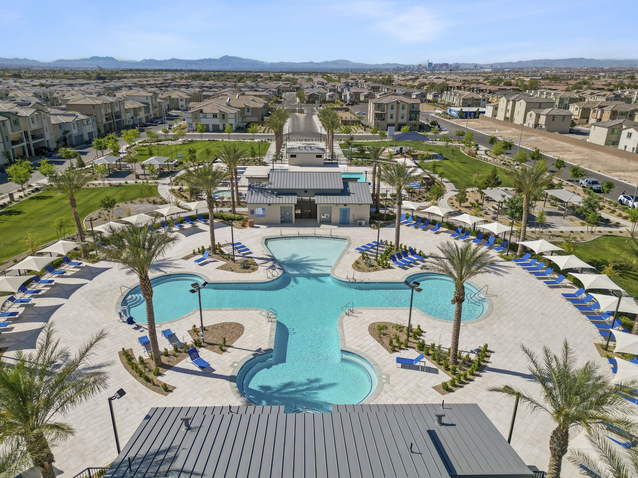 Aerial view looking down at the cross-shaped central swimming pool, clubhouses, and green parks at the Watercolor planned community, with the Las Vegas skyline in the distance.