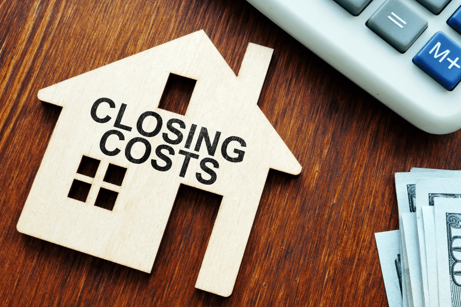 A wooden house cutout stamped with "CLOSING COSTS" sitting next to a calculator and stacked cash on a wood table for a Las Vegas townhome.