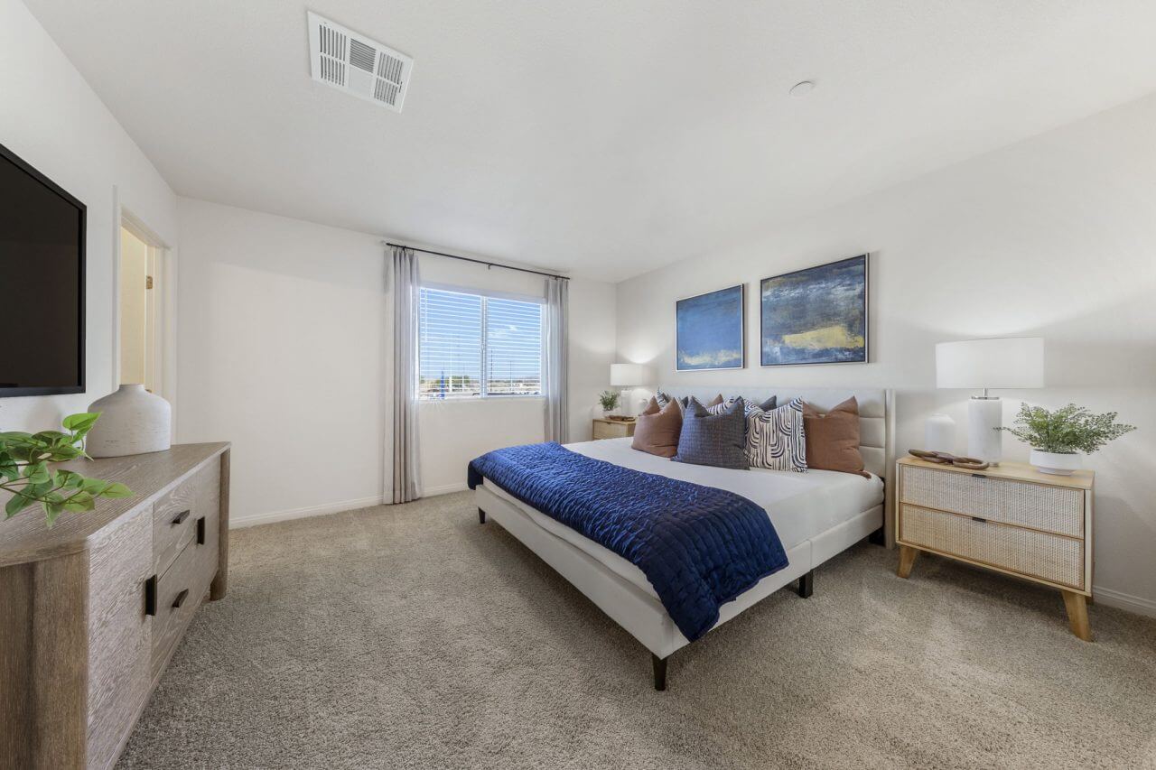 Spacious primary bedroom featuring carpeted flooring, a white-framed bed with a blue quilt, large abstract blue and gold artwork, and two woven nightstands in an Achieve 140 model home at the Independence neighborhood