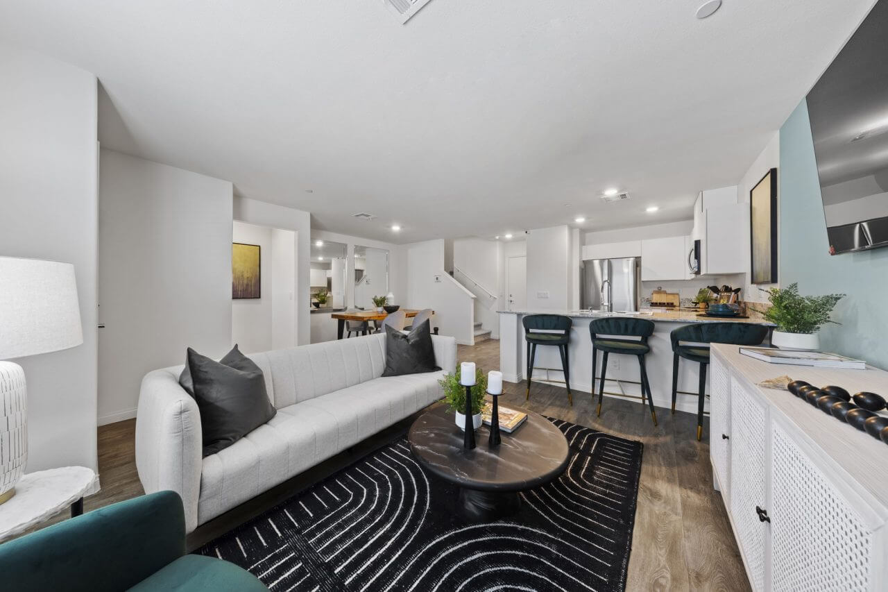 Expansive open-concept living and kitchen area featuring upgraded vinyl flooring, a white modern sofa, and a kitchen island with teal barstools in a Prosperity 200 model home at the Independence neighborhood.
