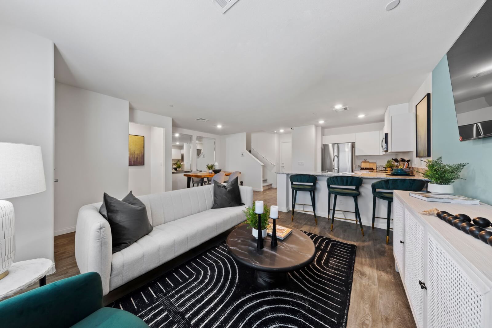 Expansive open-concept living and kitchen area featuring upgraded vinyl flooring, a white modern sofa, and a kitchen island with teal barstools in a Prosperity 200 model home at the Independence neighborhood.