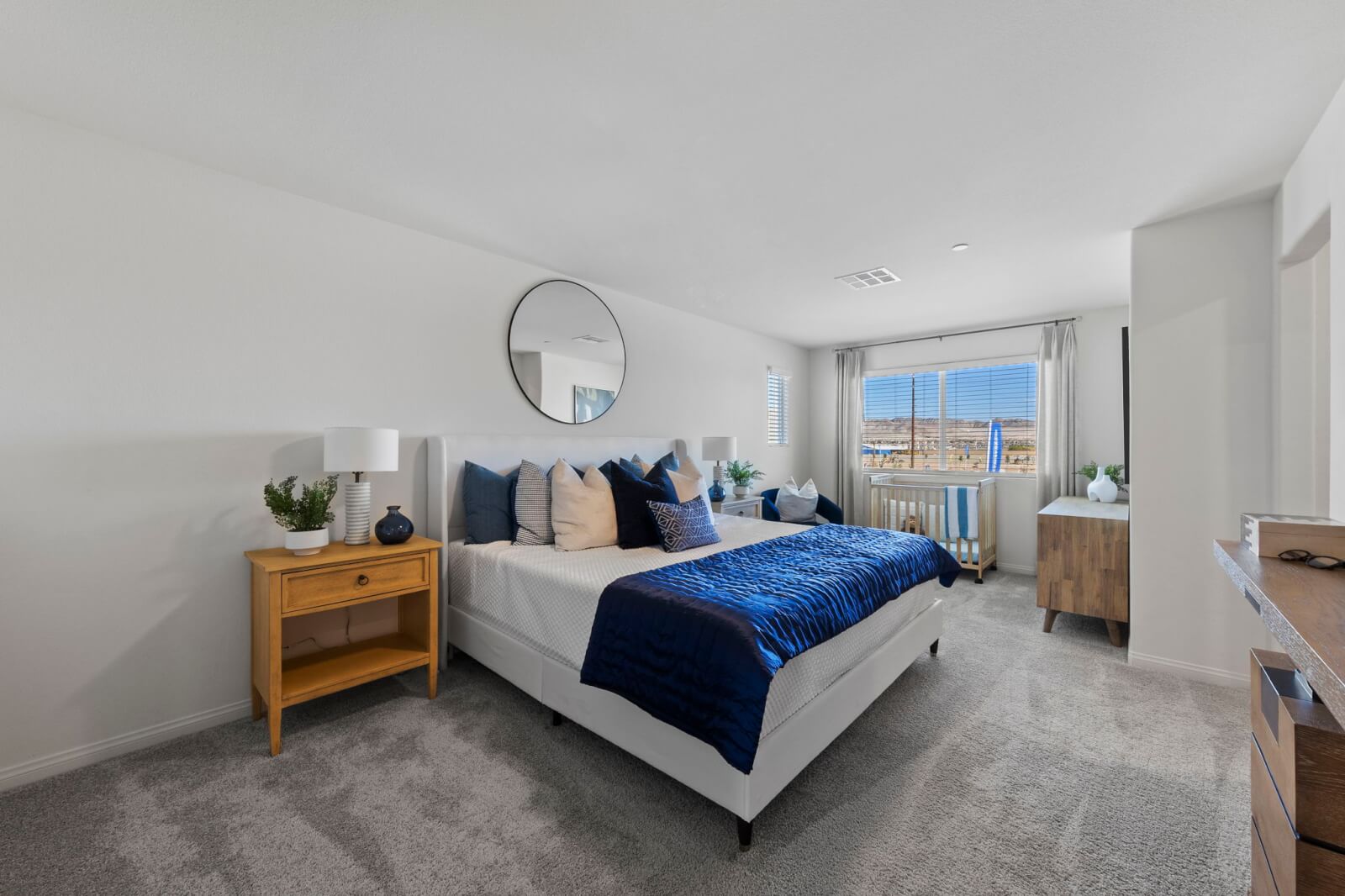 Spacious primary bedroom featuring a white upholstered bed with blue bedding, a large round mirror, and a crib near the window in a Prosperity 220 model home at the Independence neighborhood.