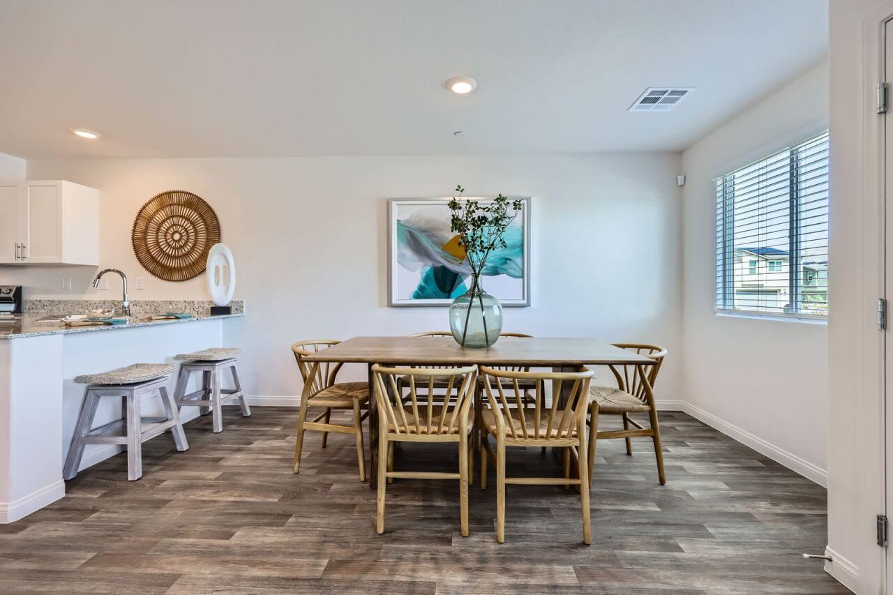 Elegant dining area with a light wood table, wishbone-style chairs, and a large abstract painting, adjacent to a modern white kitchen in a Prosperity 300 home at the