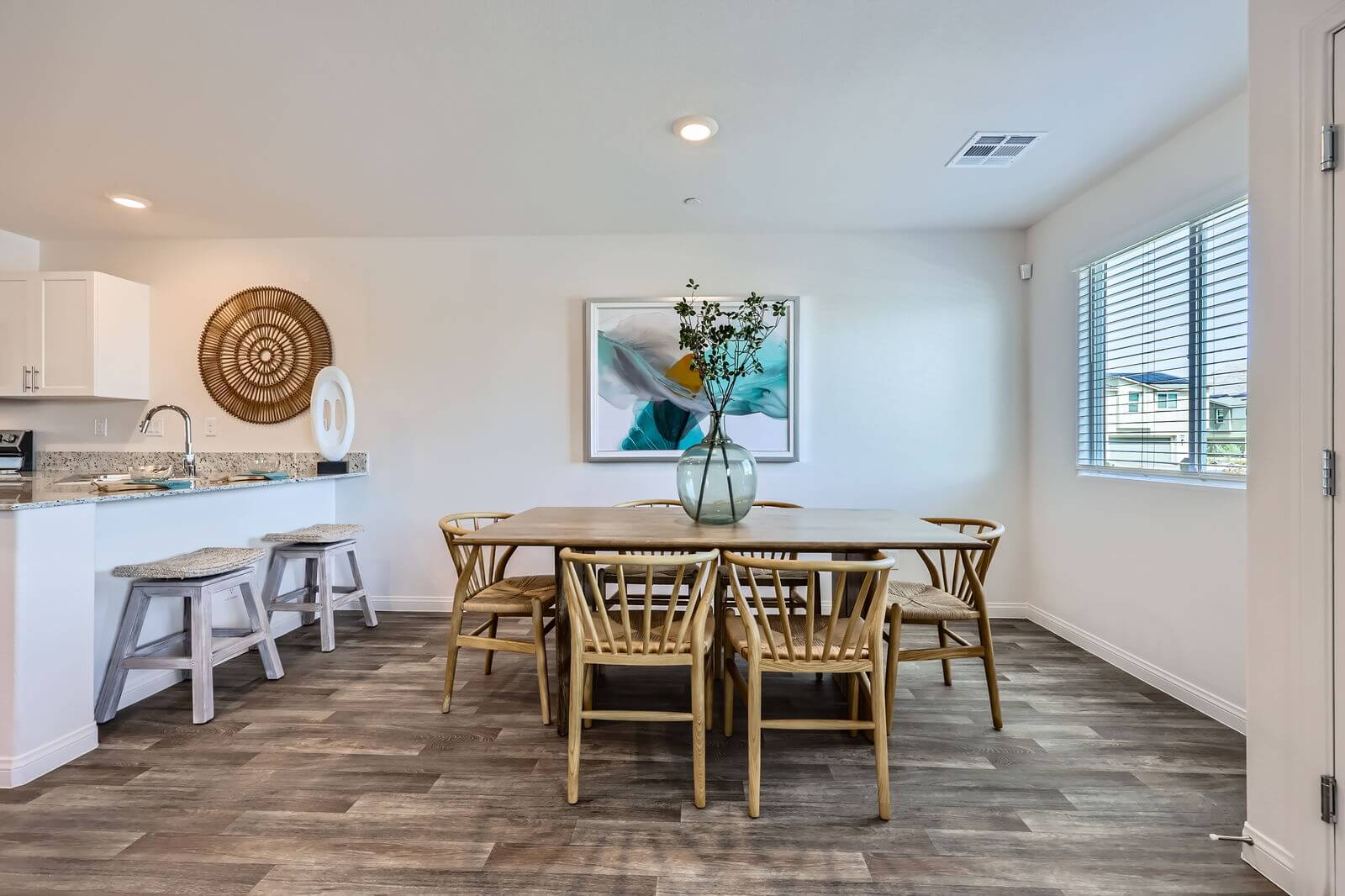 Elegant dining area with a light wood table, wishbone-style chairs, and a large abstract painting, adjacent to a modern white kitchen in a Prosperity 300 home at the