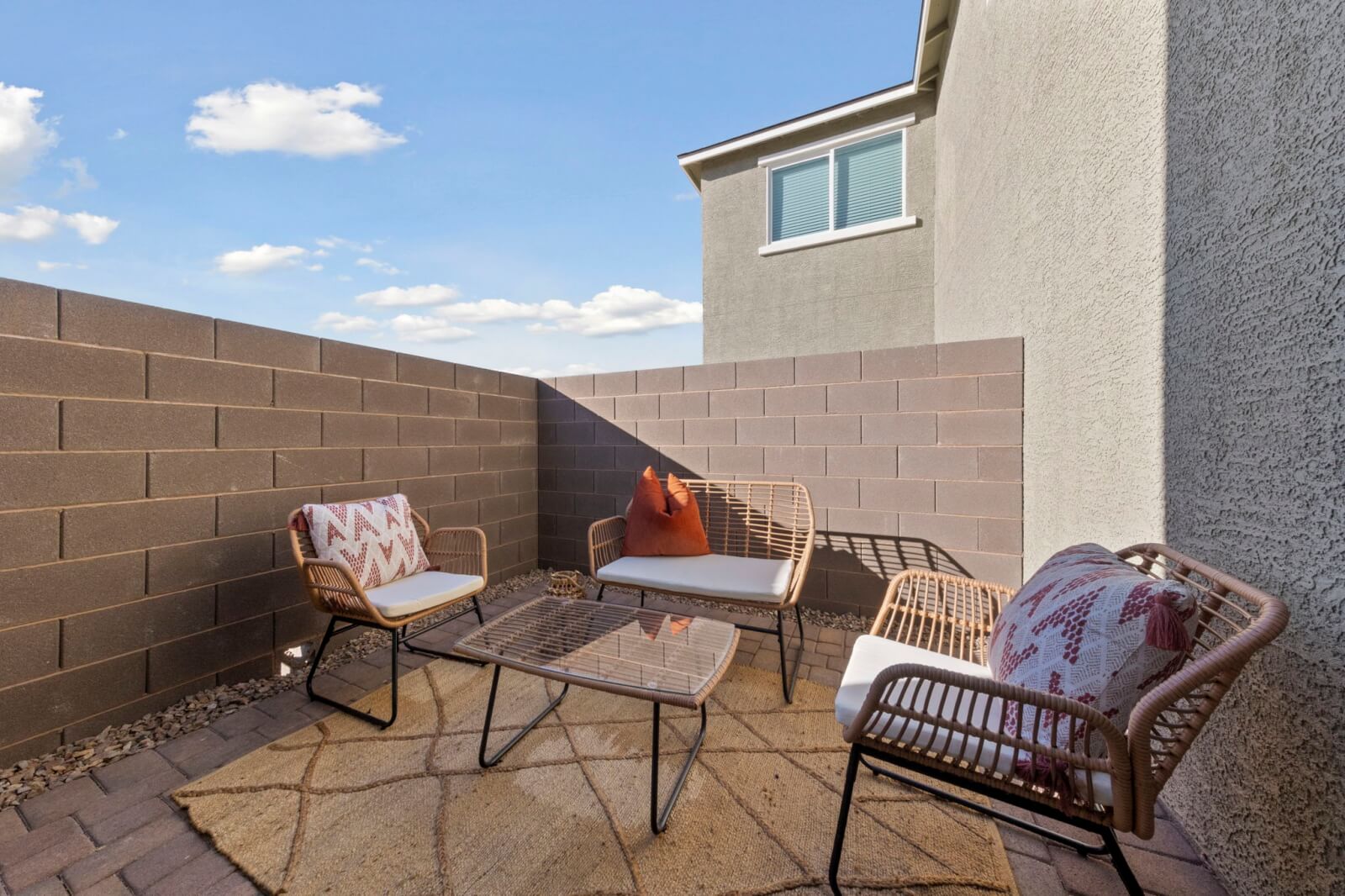 Cozy private patio area featuring brick pavers, a tan rug, and woven outdoor furniture with orange and patterned pillows at the Independence neighborhood.