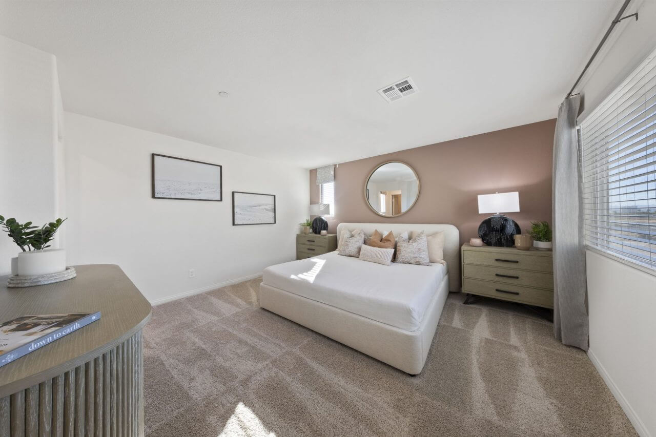 Spacious primary bedroom featuring a soft pink accent wall, a white upholstered bed, and modern furnishings in a Success 180 home at the Independence neighborhood.