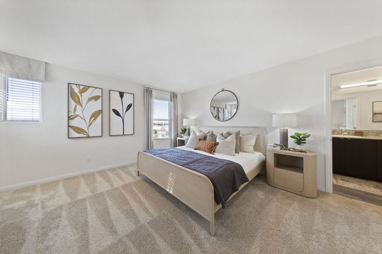 Spacious primary bedroom featuring a light wood bed, plush carpeting, and an attached en-suite bathroom in a Triumph 240 home at the Independence neighborhood.