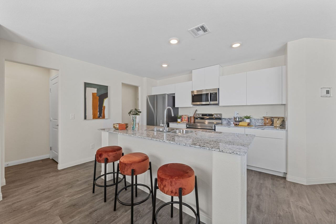 Open-concept kitchen with white cabinetry, stainless steel Samsung® appliances, and a granite island with three orange barstools at the Independence neighborhood.