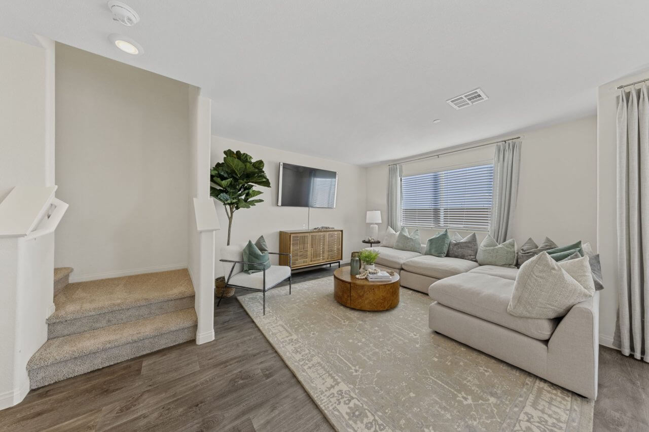 Modern living room featuring vinyl flooring, a large beige sectional sofa, and carpeted stairs leading upstairs in a Triumph 260 home at the Independence neighborhood.
