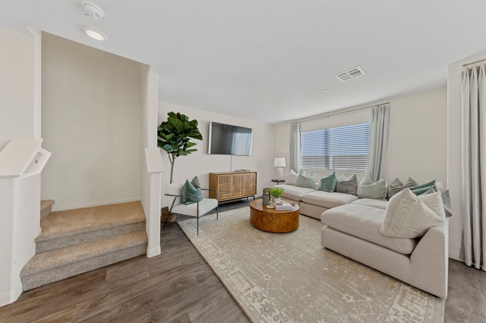 Modern living room featuring vinyl flooring, a large beige sectional sofa, and carpeted stairs leading upstairs in a Triumph 260 home at the Independence neighborhood.