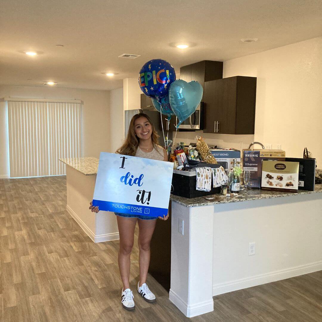 A smiling young woman celebrates buying her new Las Vegas townhome, holding an "I did it!" closing sign inside her kitchen at a Touchstone Living neighborhood.