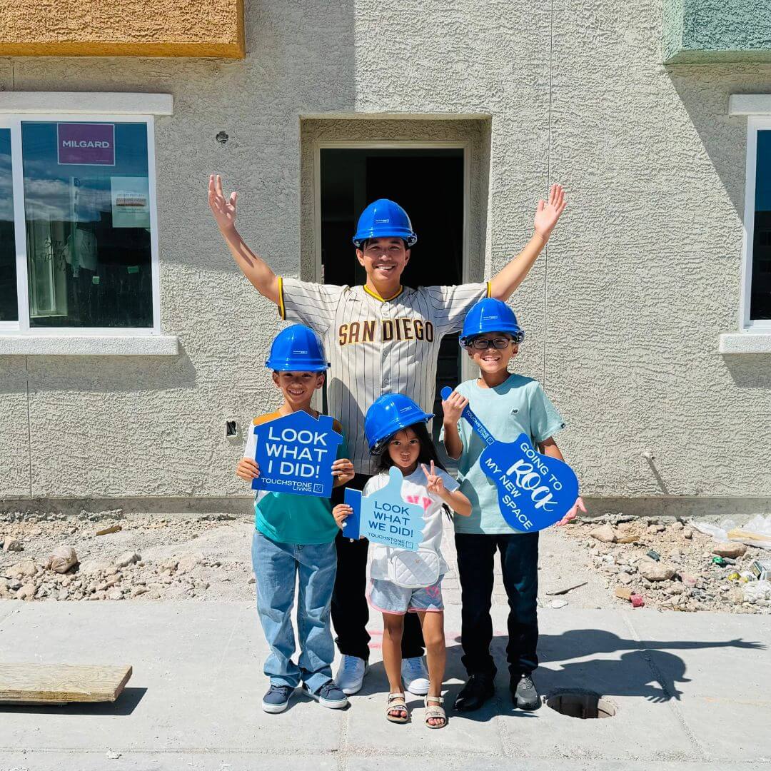 A proud father and his three children wearing blue hard hats celebrate their new Las Vegas townhome, smiling and holding closing signs outside the property at a Touchstone Living neighborhood.