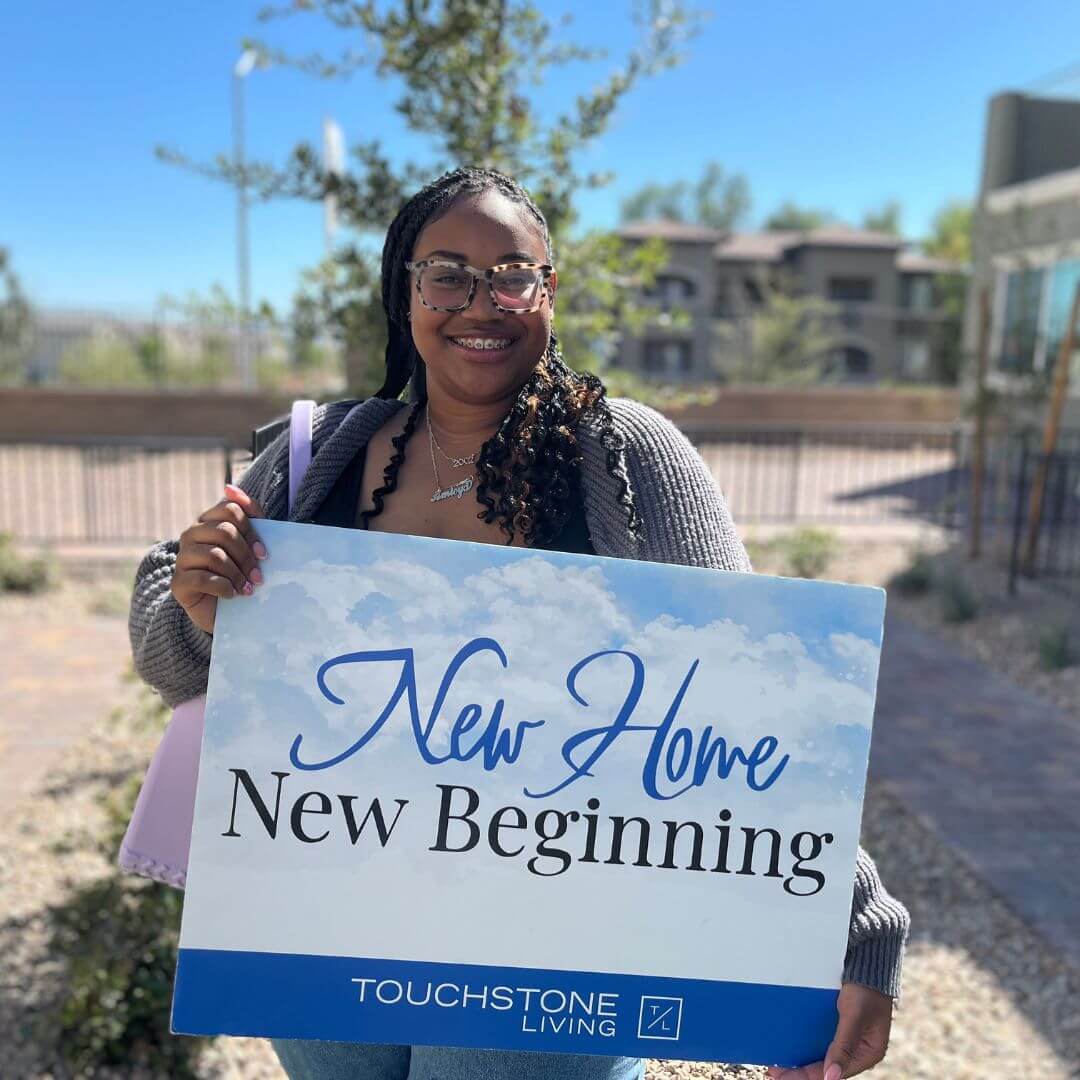 A smiling young woman celebrates purchasing her new Las Vegas townhome, holding a large "New Home, New Beginning" sign outside at a Touchstone Living neighborhood.