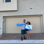 A young couple celebrates buying their new Las Vegas townhome, smiling and holding up closing signs on a brick paver driveway at a Touchstone Living neighborhood.