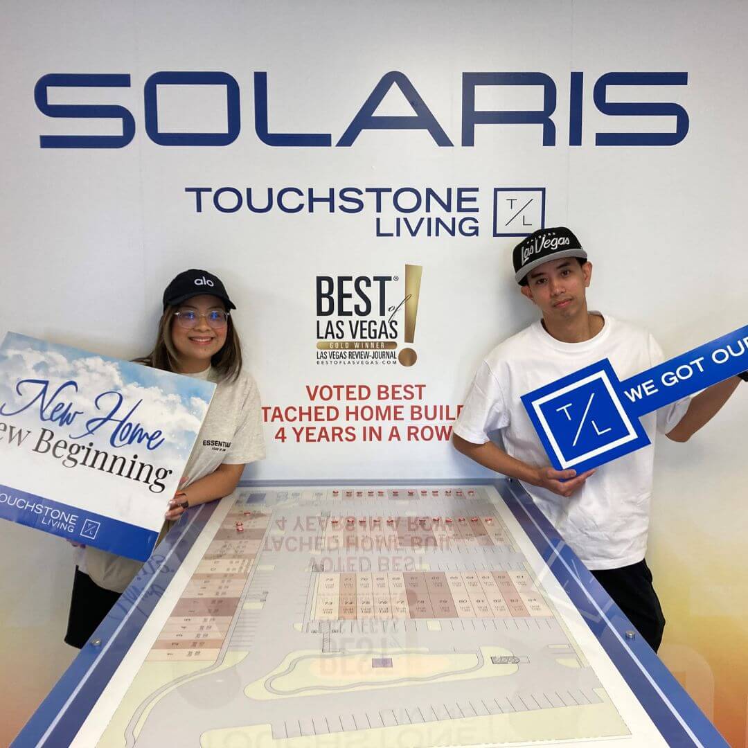 Happy new homeowners smiling and holding closing signs in front of a Solaris sign at a Touchstone Living neighborhood in Las Vegas.