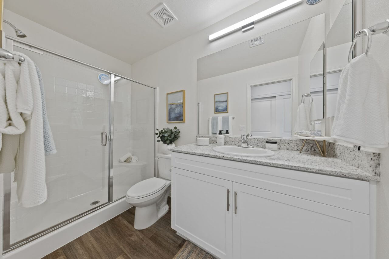 Modern full bathroom featuring a white vanity with granite countertops, a glass-enclosed shower, and upgraded vinyl flooring at the Solaris at Warm Springs neighborhood.