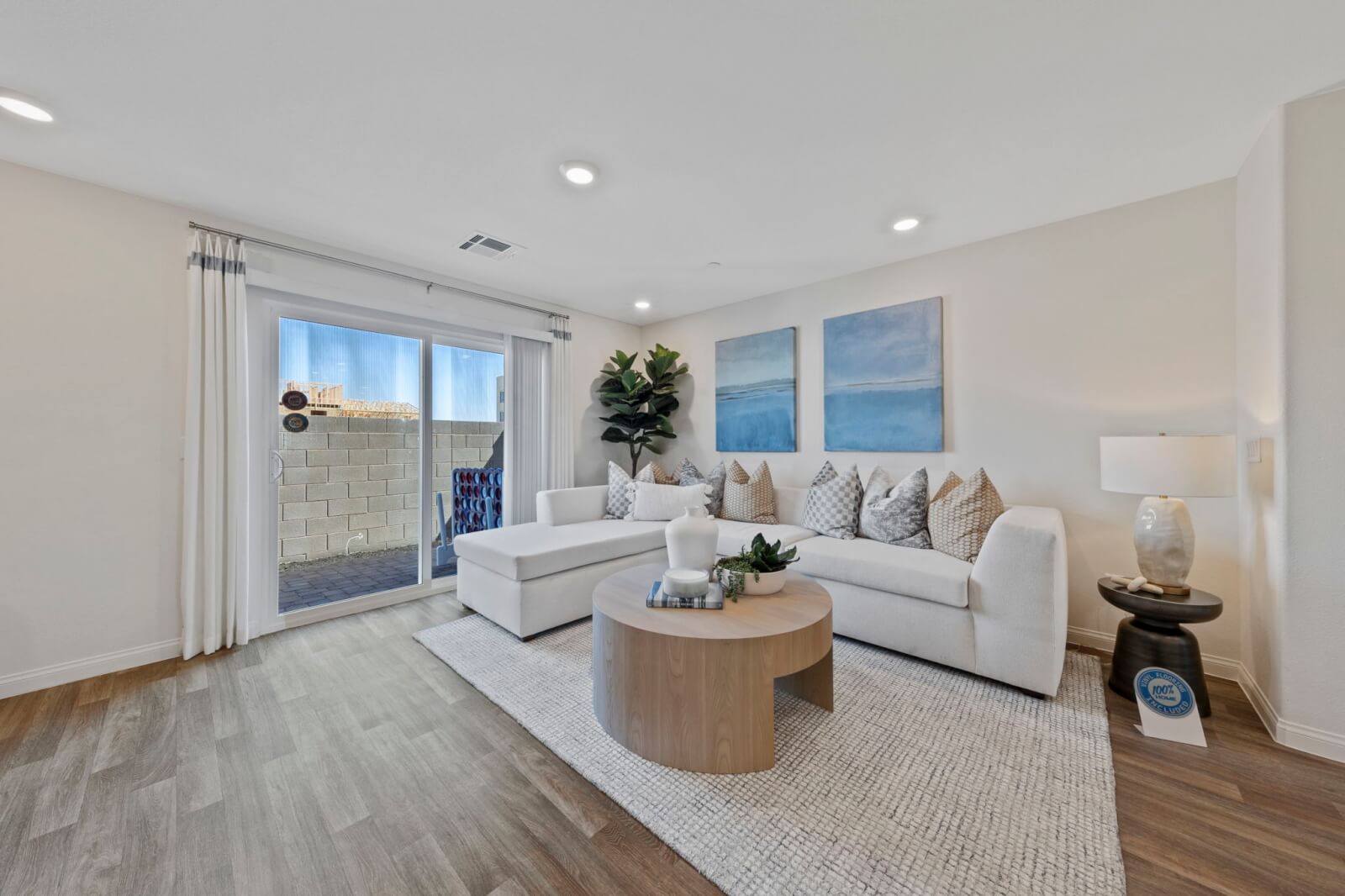 Bright living room featuring upgraded vinyl flooring, a white sectional sofa, a round wood coffee table, and blue abstract art in a Solaris model at the Solaris at Warm Springs neighborhood.