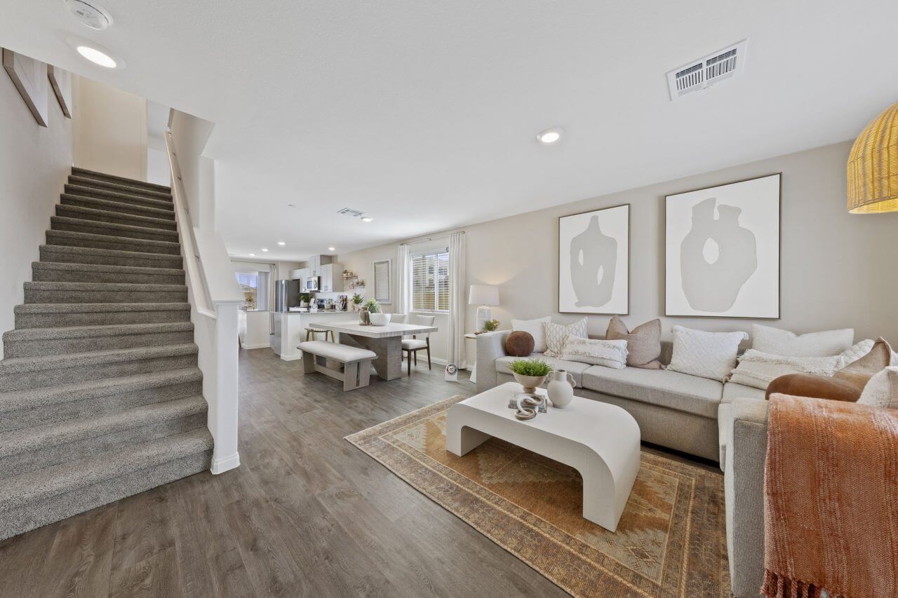 Open-concept living room and dining area featuring upgraded vinyl flooring, a light beige sectional sofa, and carpeted stairs in plans 100 & 130 at the Solaris at Warm Springs neighborhood.