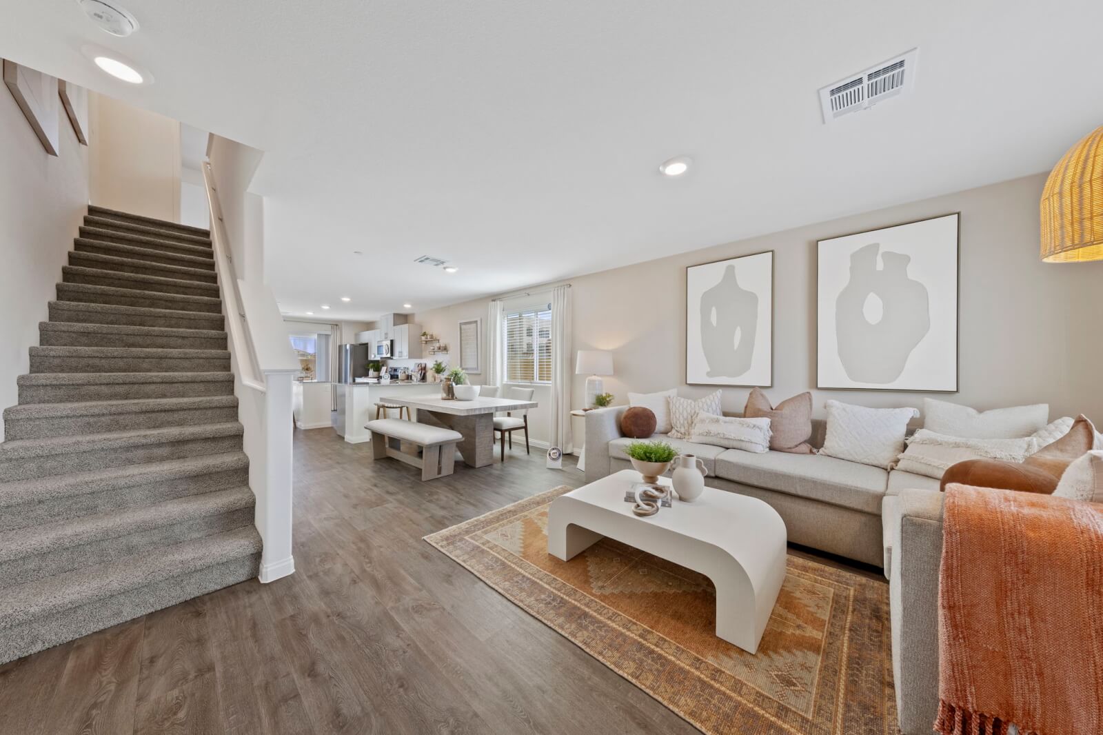 Open-concept living room and dining area featuring upgraded vinyl flooring, a light beige sectional sofa, and carpeted stairs in plans 100 & 130 at the Solaris at Warm Springs neighborhood.
