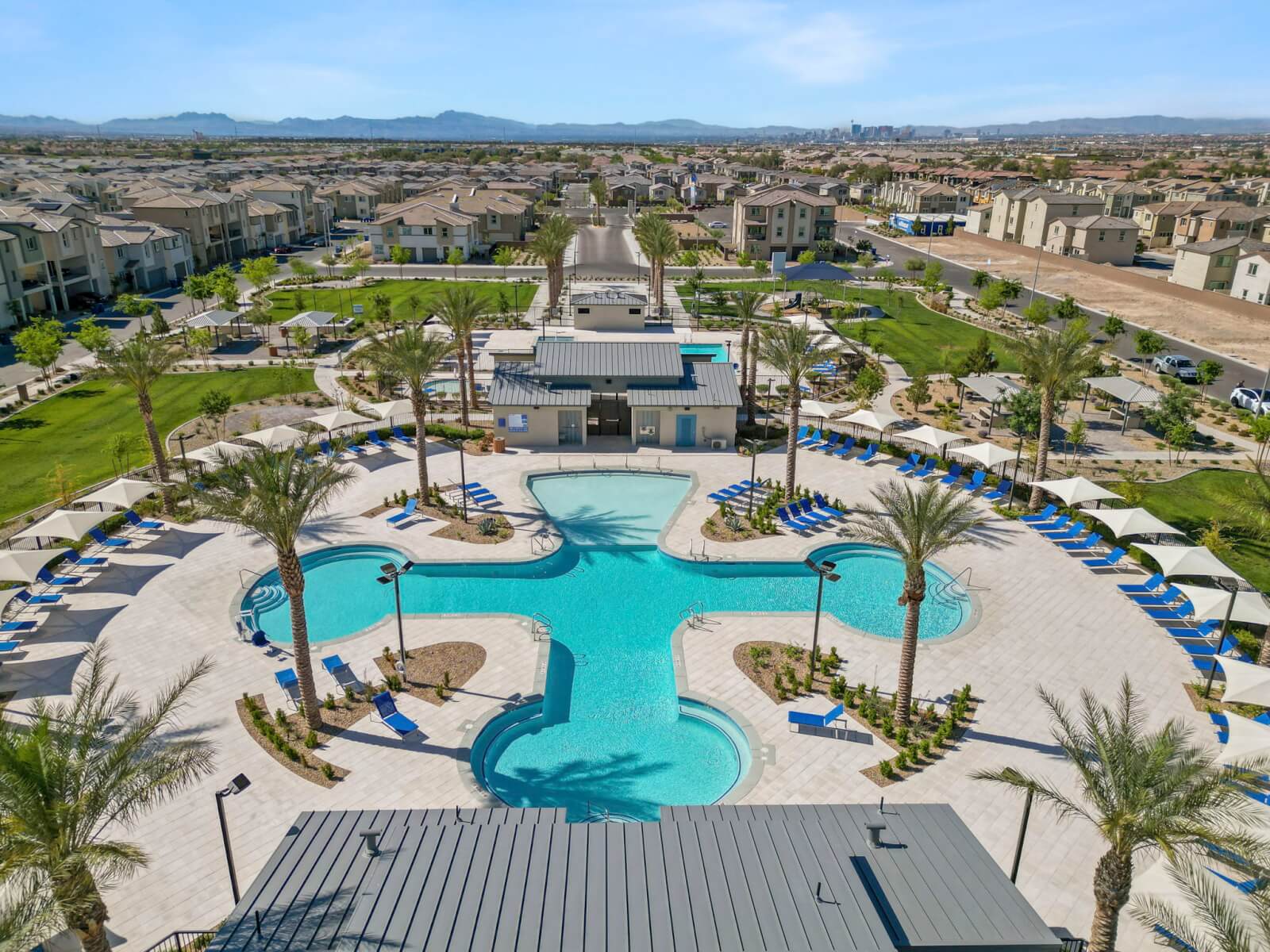 Aerial view looking down at the cross-shaped central swimming pool, clubhouses, and green parks at the Watercolor planned community, with the Las Vegas skyline in the distance.