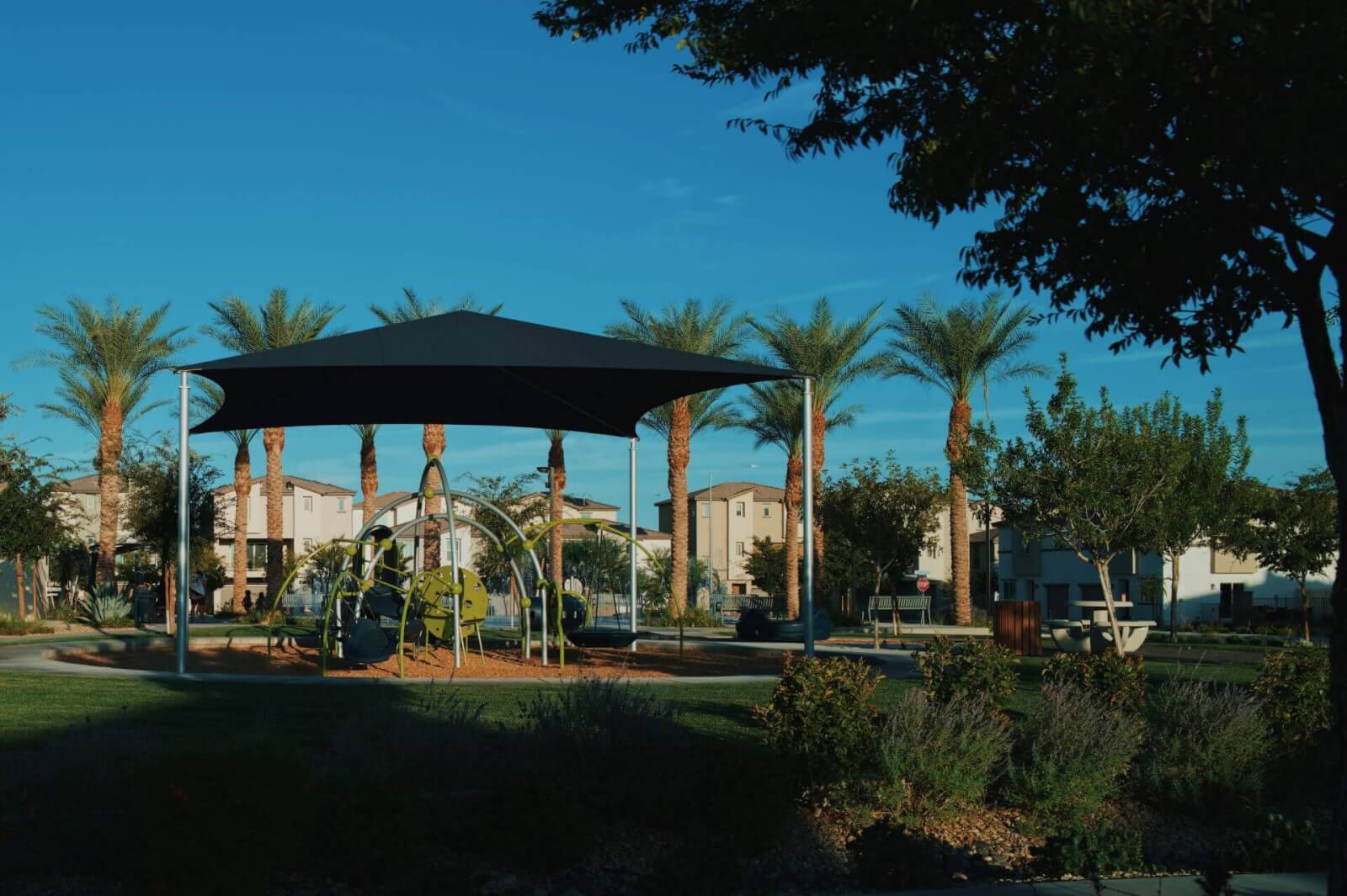 A modern playground featuring a large black sunshade canopy and climbing equipment surrounded by green grass and palm trees at the Watercolor neighborhood.