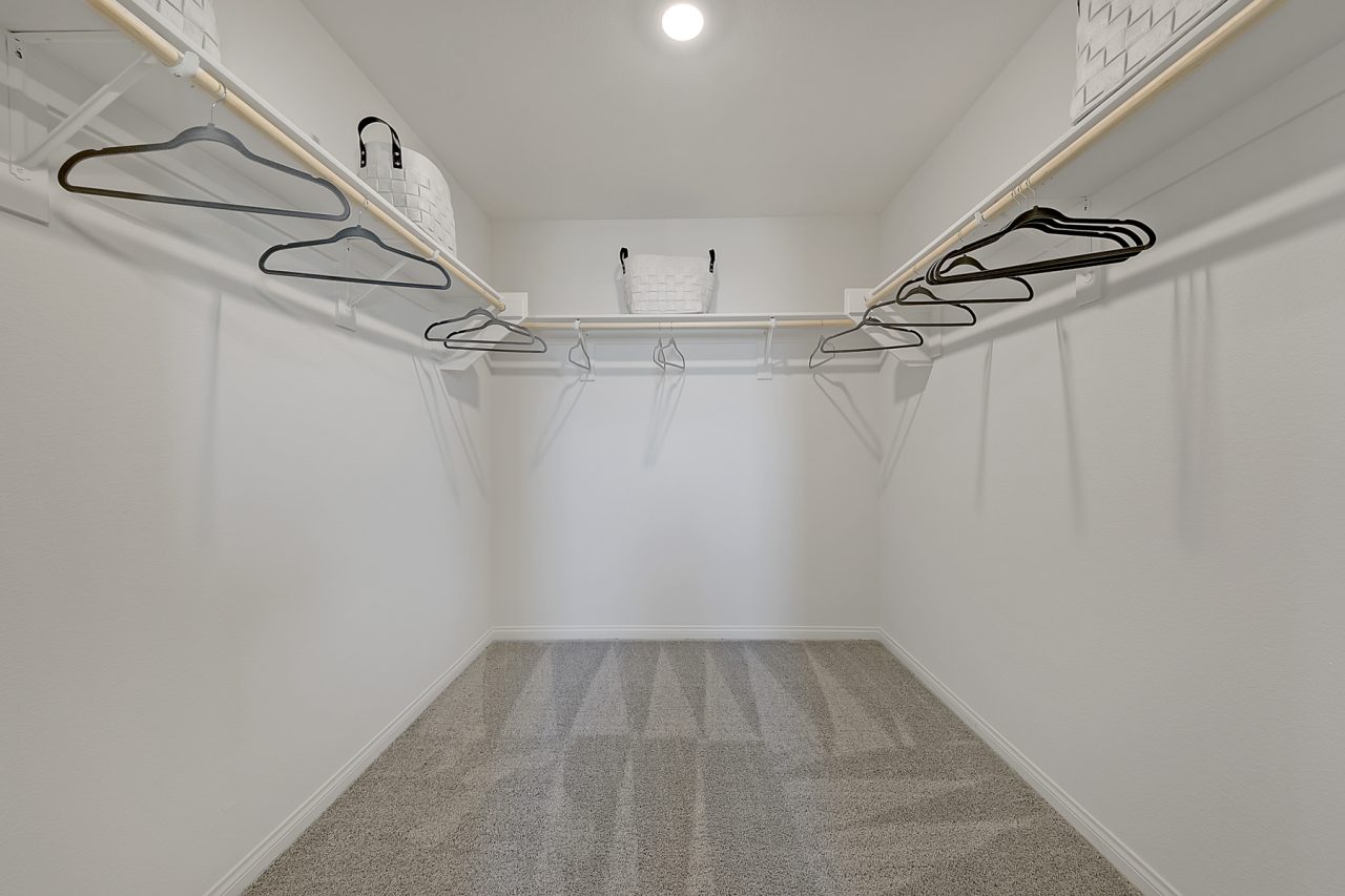 Spacious walk-in closet featuring beige carpeting, white built-in shelving, and wooden clothing rods in a Crimson Plan 520 model home.