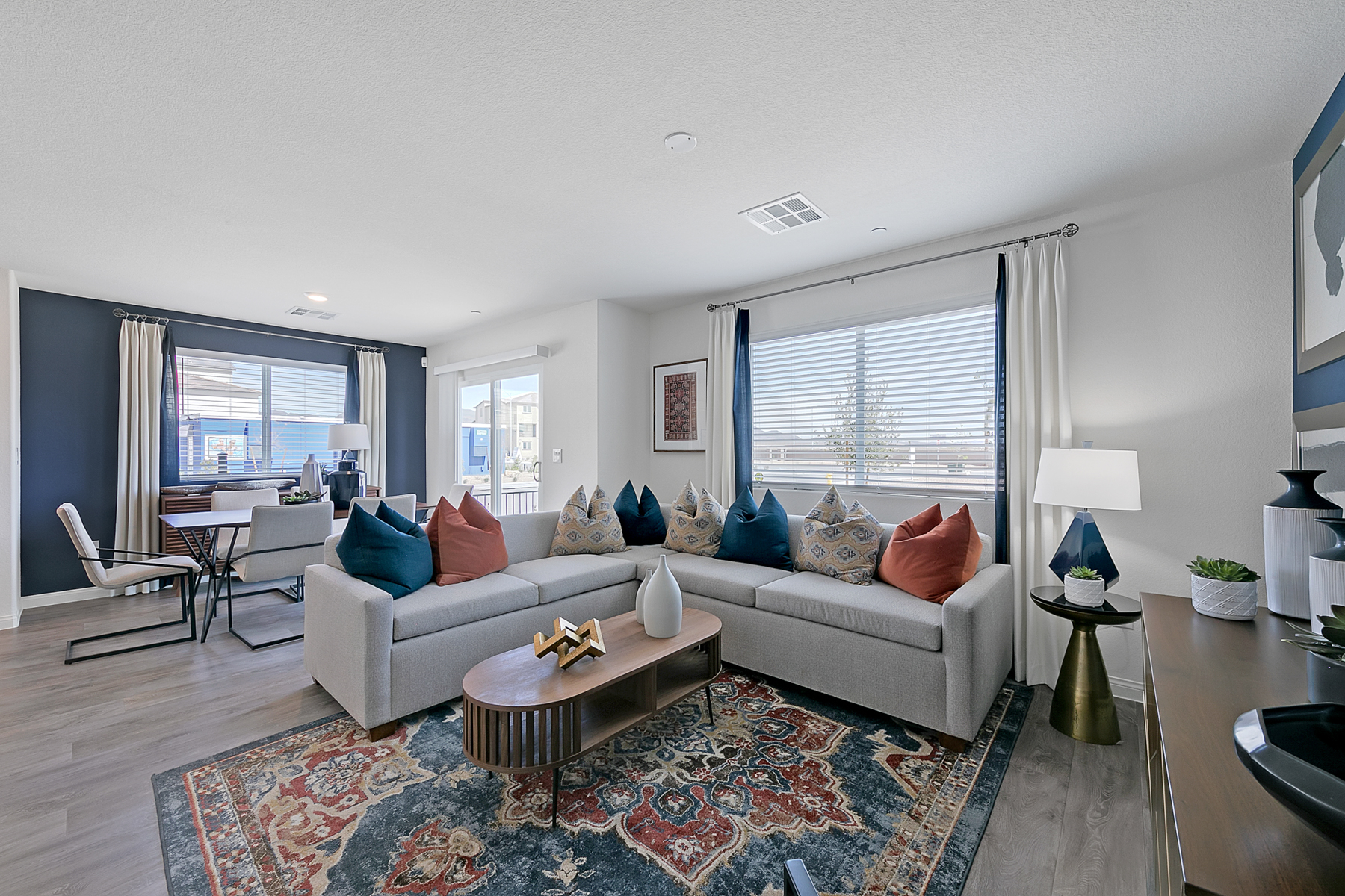 Open-concept living room featuring a grey sectional, upgraded vinyl flooring, a wood coffee table, and a dining area against a dark blue wall in a Crimson Plan 520 model home.
