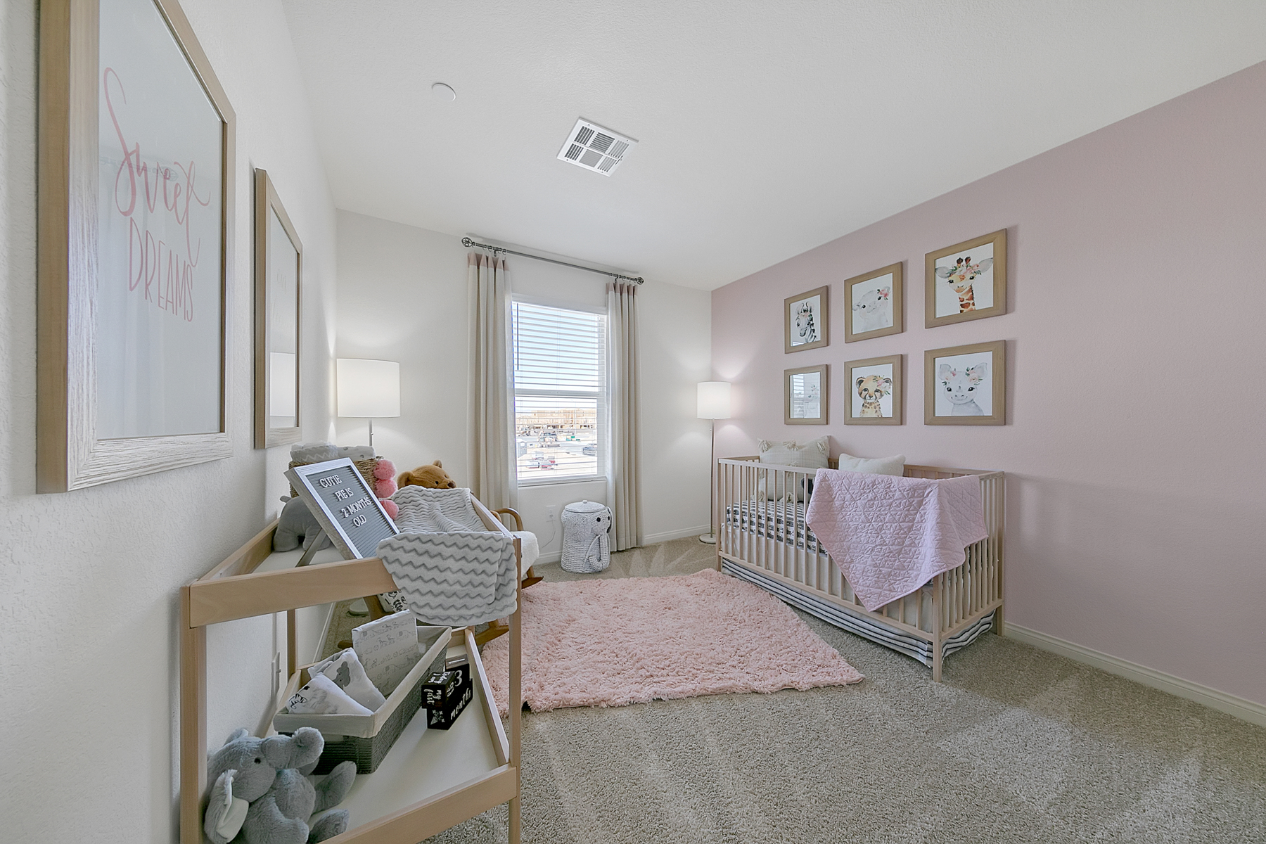 Secondary bedroom staged as a nursery with a light wood crib, a pink accent wall, animal art, and a pink rug in a Crimson Plan 523 model home.
