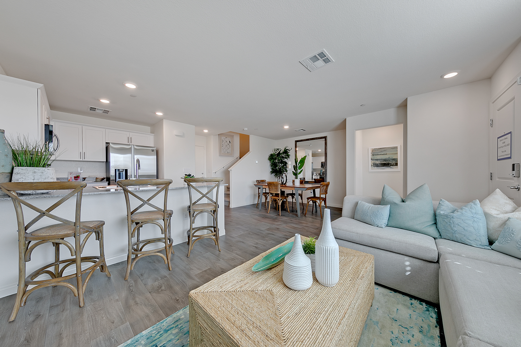 Living room featuring upgraded vinyl flooring, a light grey sectional sofa, a woven coffee table, and a view into the kitchen in a Crimson Plan 524 model home.