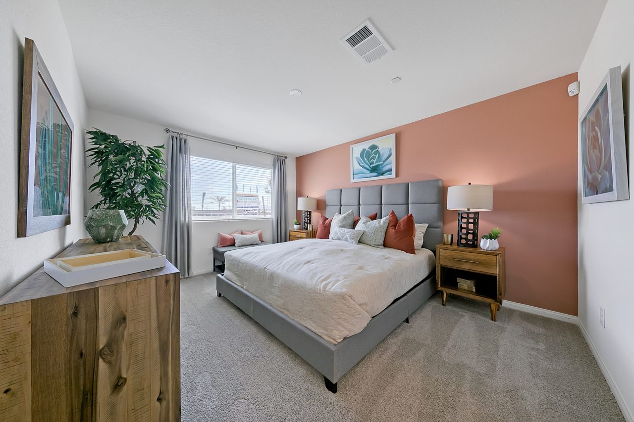 Primary bedroom featuring a coral accent wall, grey upholstered bed, wood dresser, and a faux plant in a Crimson Plan 525 model home.