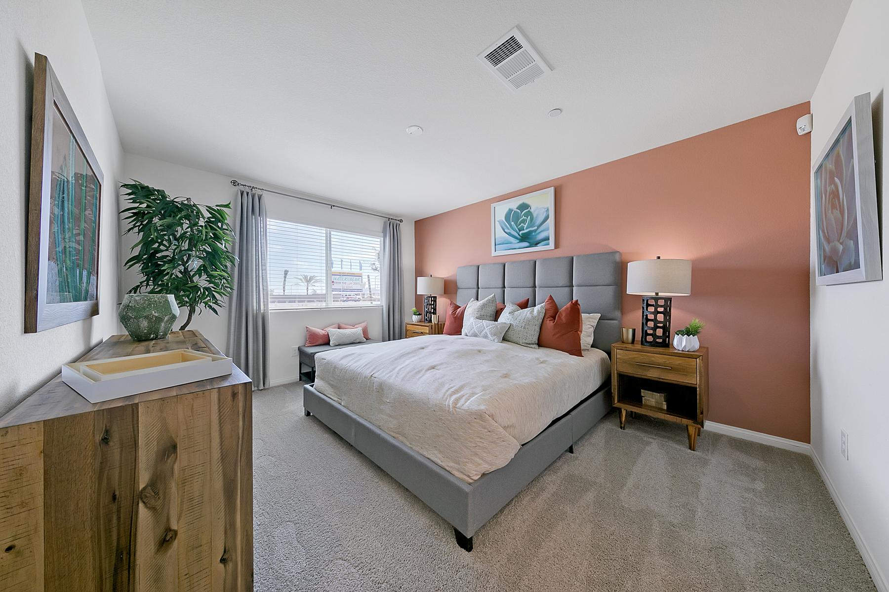 Primary bedroom featuring a coral accent wall, grey upholstered bed, wood dresser, and a faux plant in a Crimson Plan 525 model home.