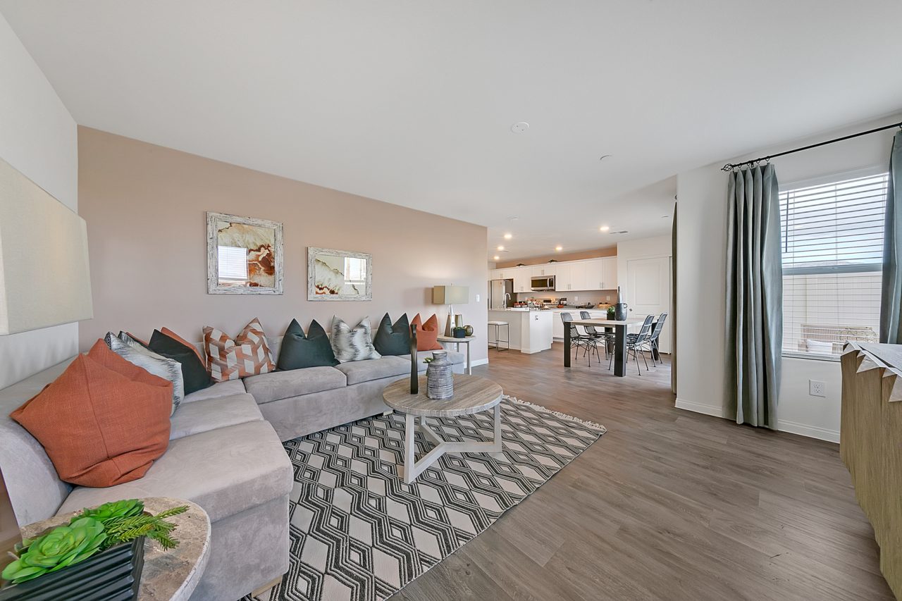 Open-concept living room featuring upgraded vinyl flooring, a grey sectional, and a view into the kitchen with granite countertops in an Indigo Plan 542 model home.