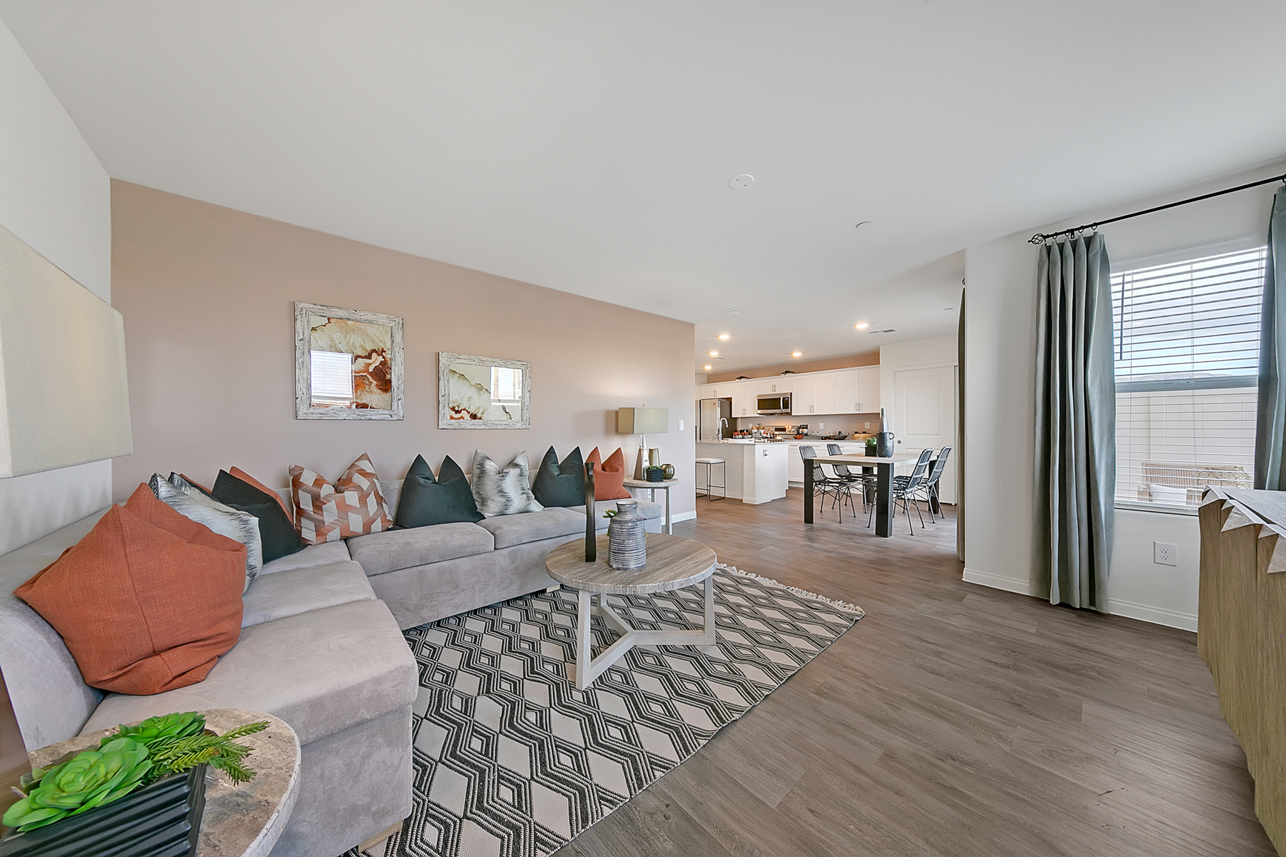 Open-concept living room featuring upgraded vinyl flooring, a grey sectional, and a view into the kitchen with granite countertops in an Indigo Plan 542 model home.