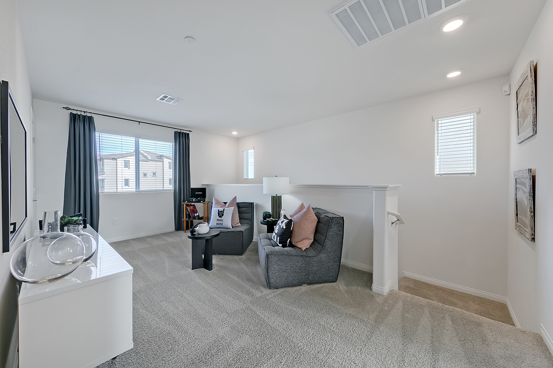 Carpeted loft space featuring two dark grey lounge chairs, a white media console, and a half-wall overlooking the stairs in an Indigo Plan 542 model home.