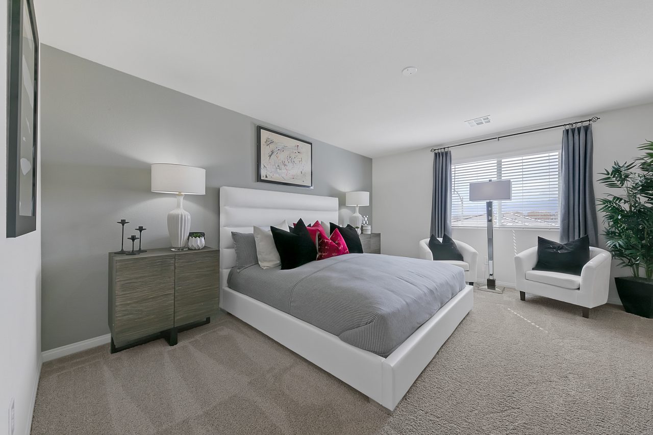 Primary bedroom featuring a white upholstered bed, grey dresser, two white armchairs, and a grey accent wall in an Indigo Plan 543 model home.