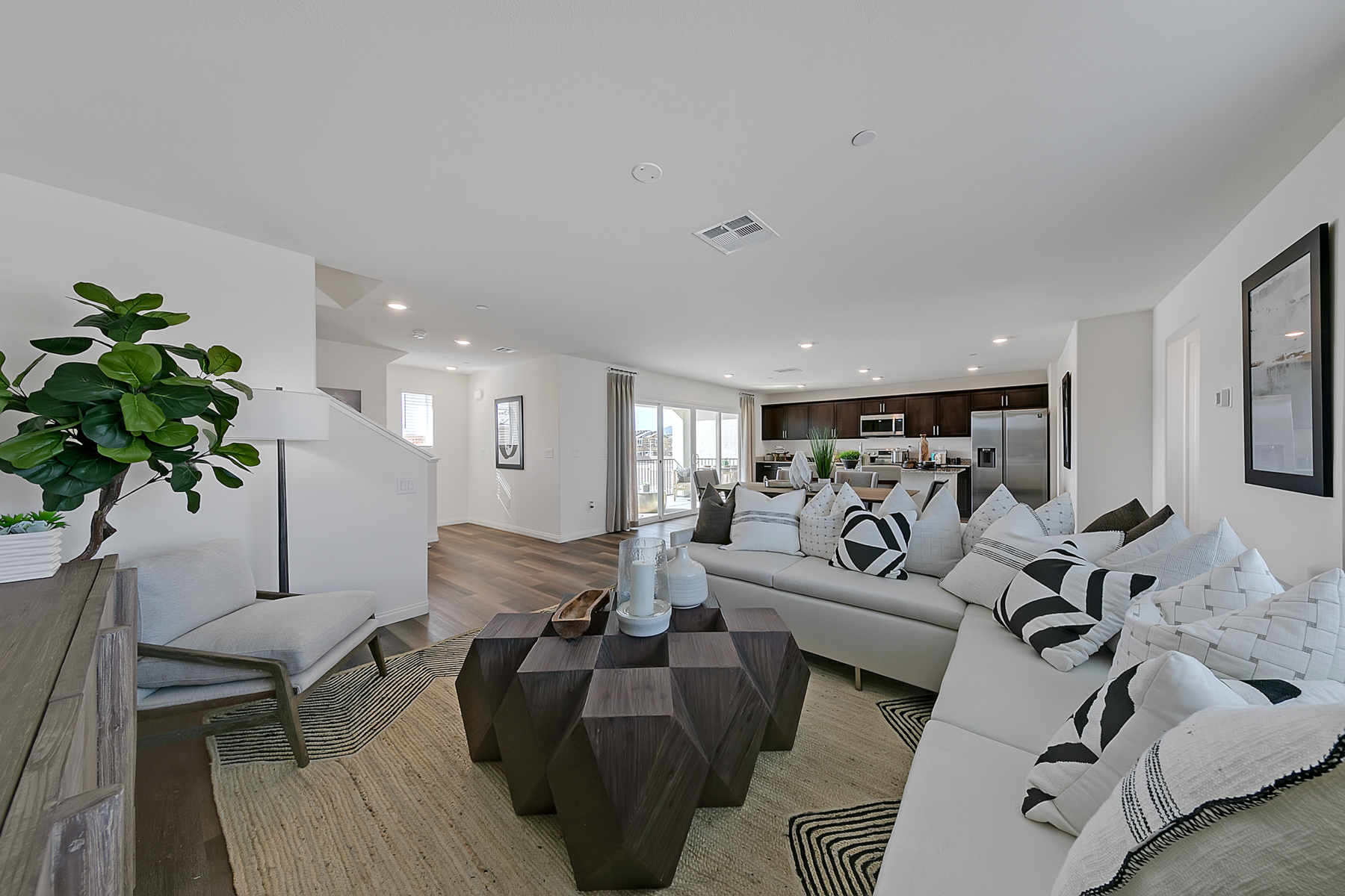 Open-concept living room featuring upgraded vinyl flooring, a white sectional sofa, a geometric coffee table, and a view of the kitchen in an Indigo Plan 544 model home.