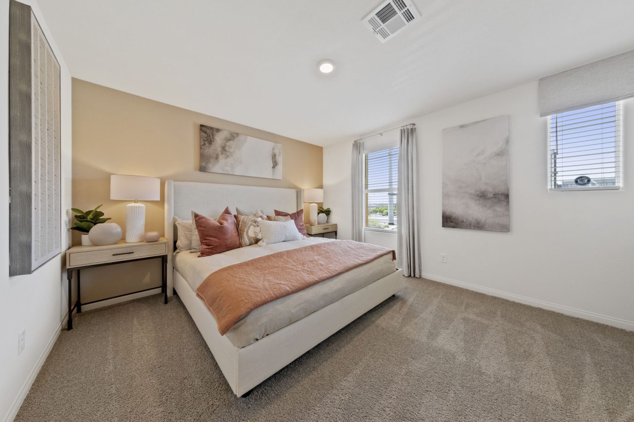 Primary bedroom with carpet and a soft peach accent wall featuring a wood-frame bed with neutral and rust bedding and a geometric mirror in a Solaris planned community home.