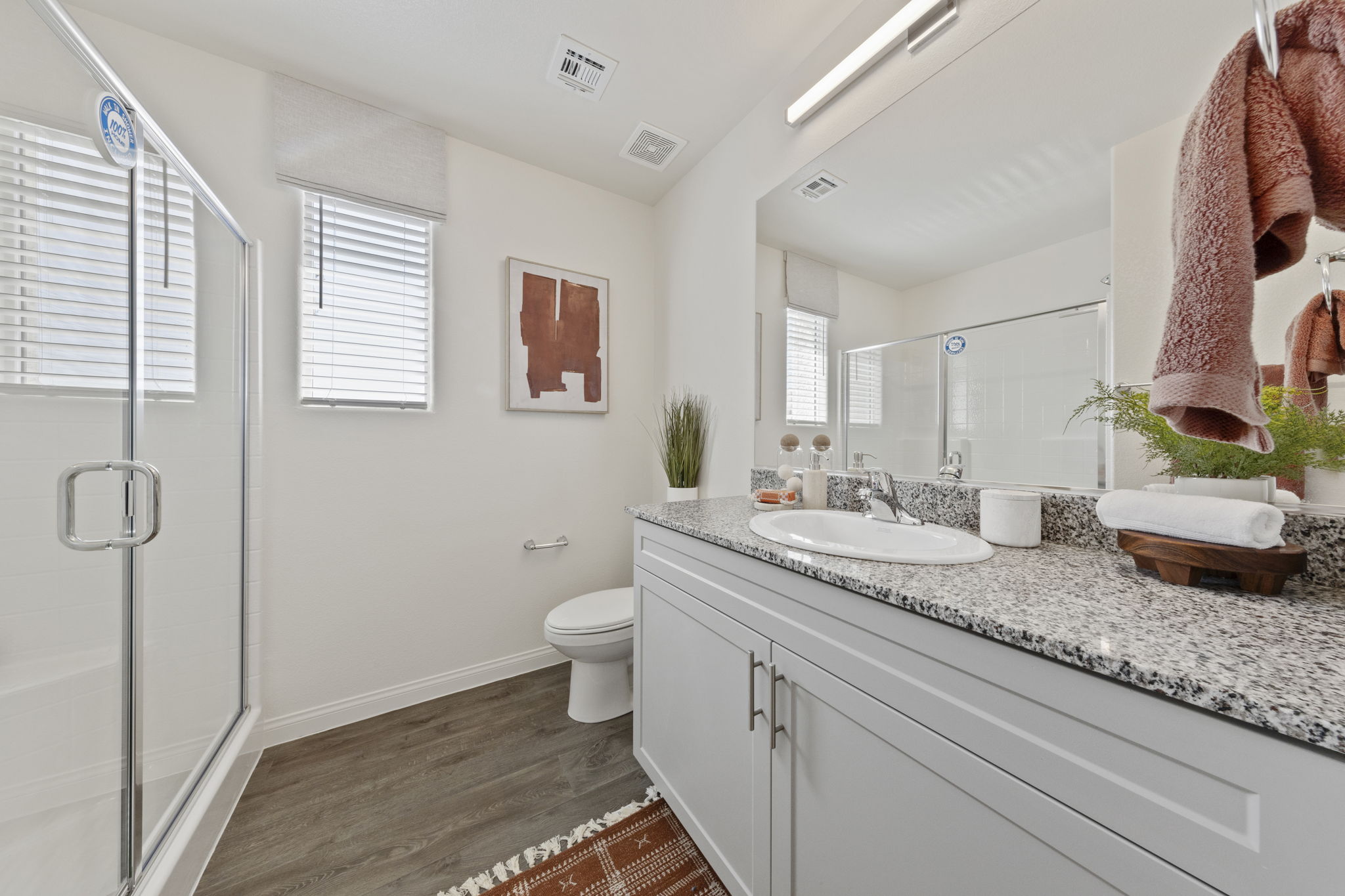 Luxurious primary bathroom with dark wood-look upgraded vinyl plank flooring, a grey granite double vanity, a walk-in glass shower, and a soaking tub under a window.